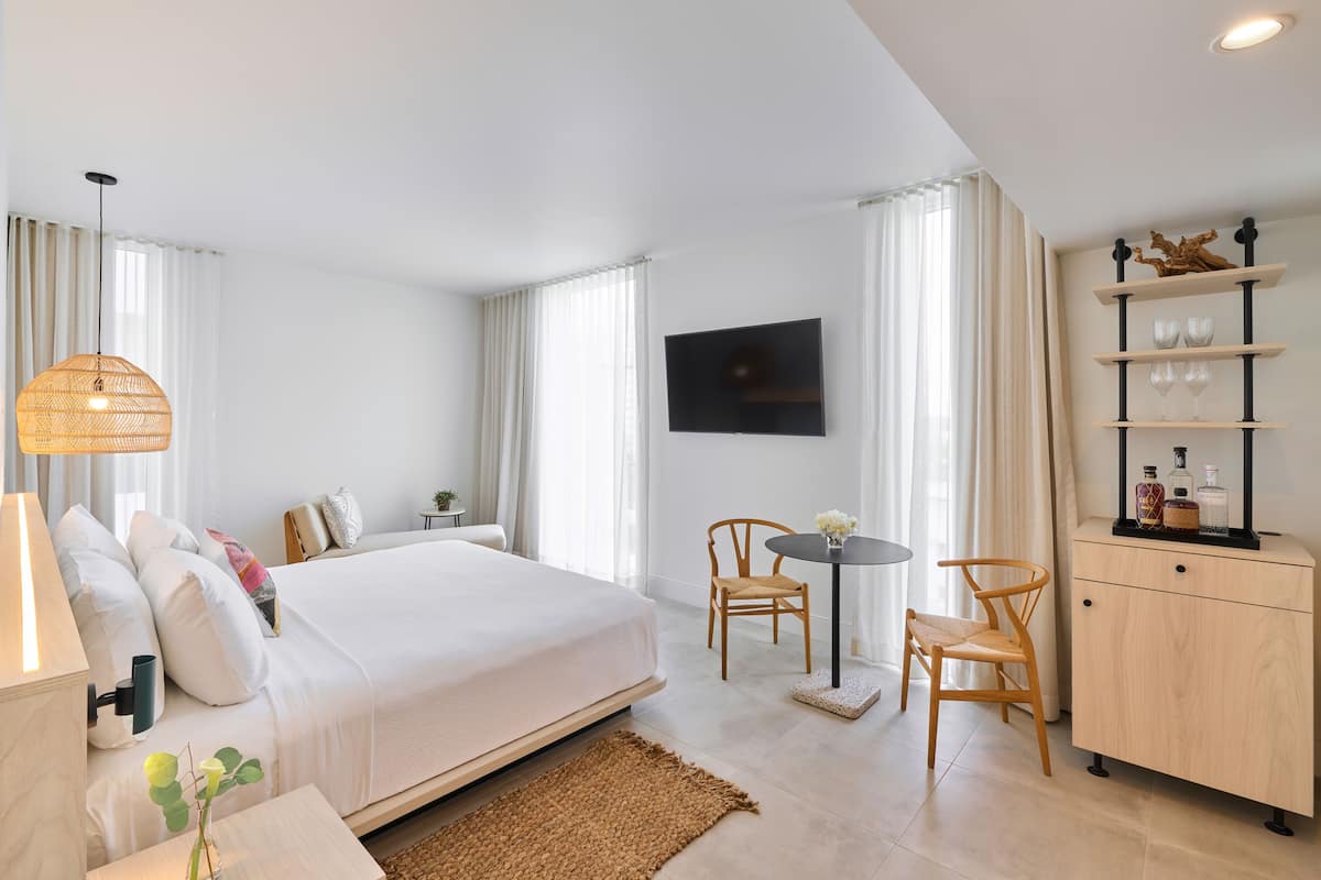 A hotel room with a bed, bedside table, lamp, chair, small table, and a cabinet with shelves.