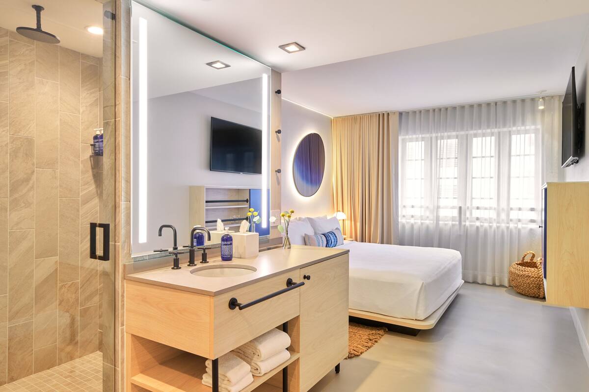 A modern hotel room with a large mirror, a double bed, a bedside table, a TV mounted on the wall, and a bathroom with a shower and sink.