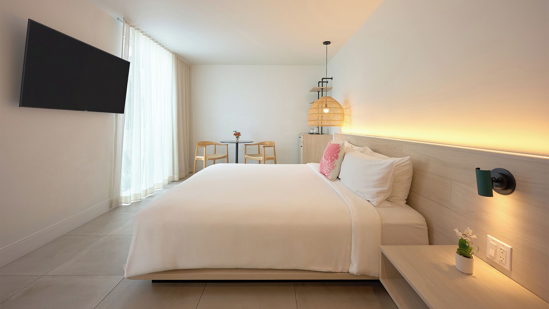 A modern hotel room with a large bed, a flat-screen TV mounted on the wall, a small table with chairs, and a wall-mounted lamp.