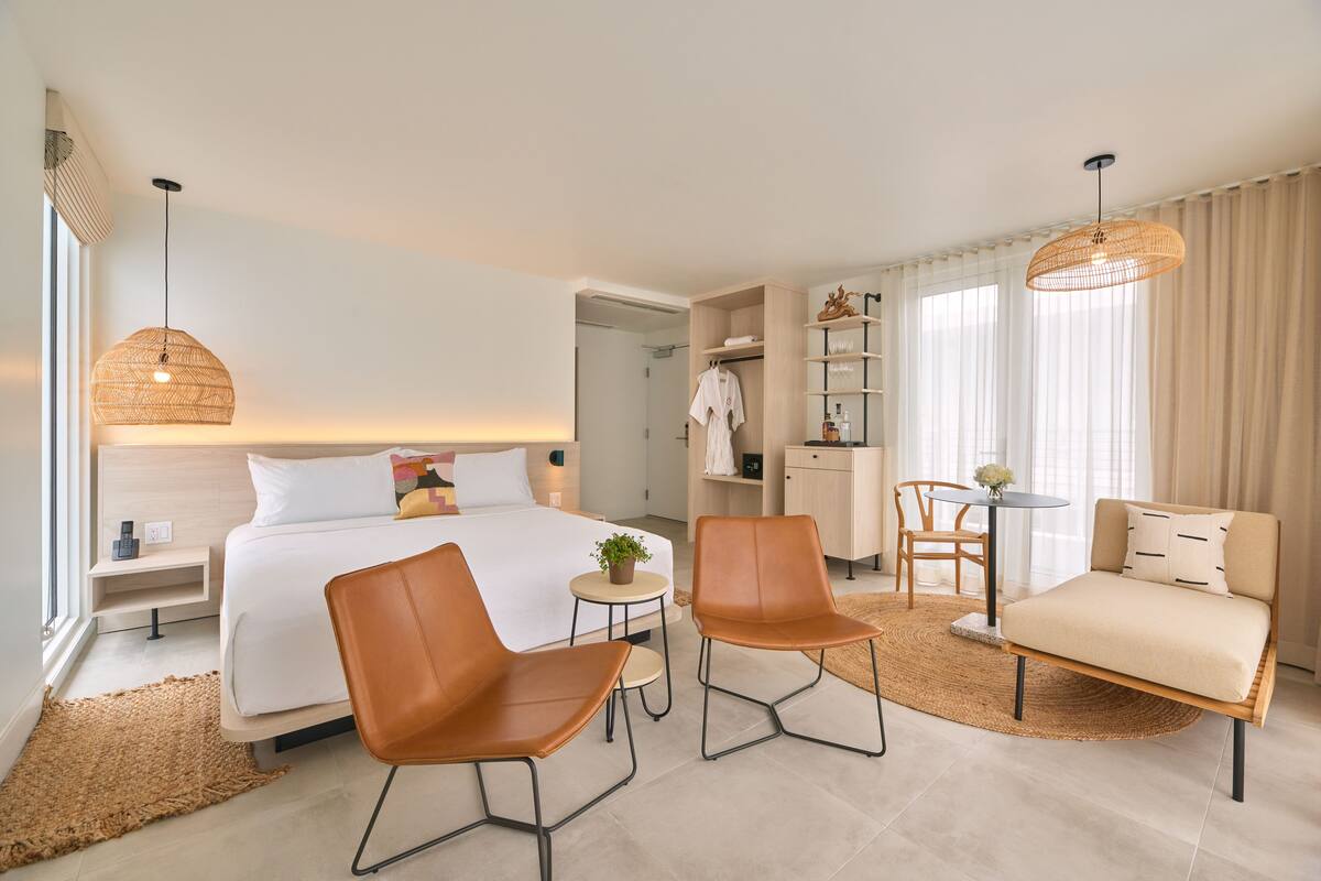 A modern hotel room with a bed, two brown chairs, a small table, a sofa, a wardrobe, and a hanging light fixture.