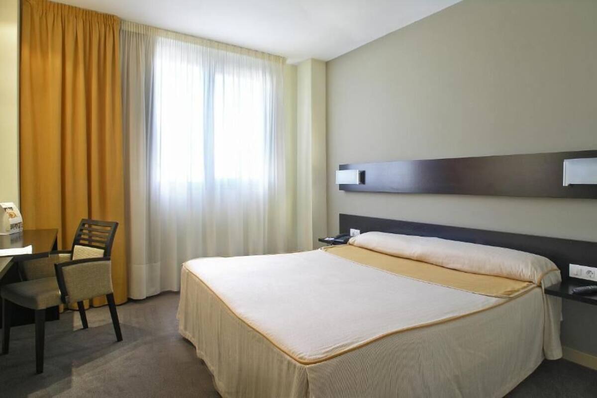 A hotel room with a large bed, a desk, a chair, and a window with curtains.