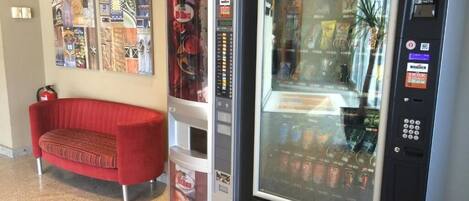 A vending machine with a glass display, a red sofa, and a wall with various posters.