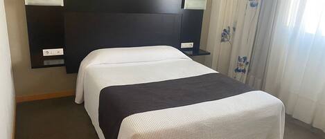 Habitación de hotel con cama, cabecera, mesita de noche y ventana con cortinas.