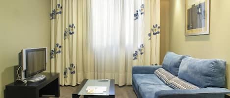 Ein Wohnzimmer mit einer Couch, einem Couchtisch, einem Fernseher auf einem Schrank und Vorhängen mit einem Blumenmuster.