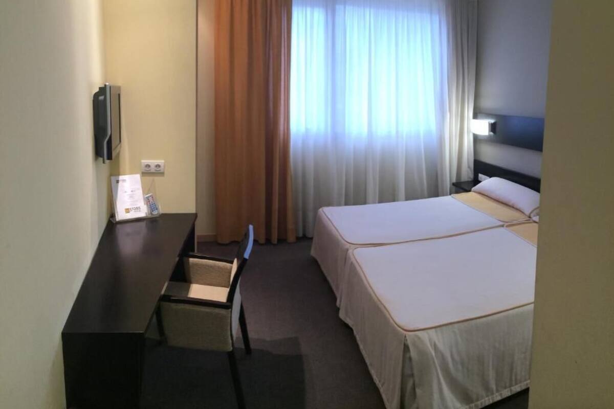 A hotel room with a desk, two beds, a window with curtains, and a wall-mounted TV.