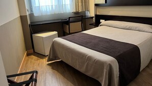 Una habitación de hotel con cama, escritorio, silla y ventana con cortinas.