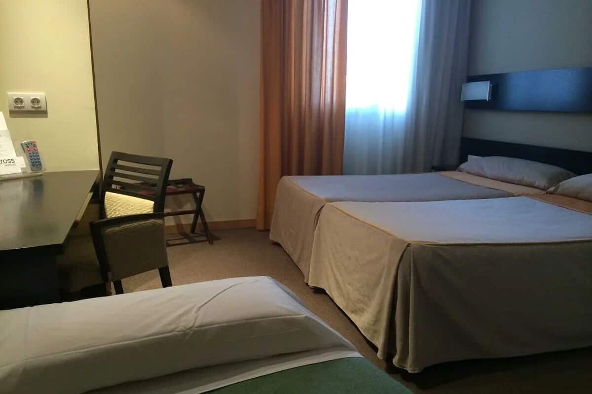 A hotel room with two single beds, a desk with a chair, and a window with curtains.