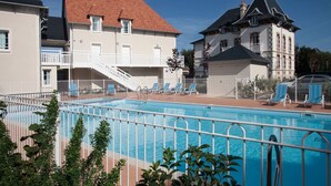 A swimming pool with a white fence, lounge chairs, and a building with a red roof and balconies. - Apartment in France with Balcony or Terrace (Cabourg)
