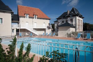A swimming pool with a white fence, lounge chairs, and a building with a red roof and balconies. - Apartment in France with Balcony or Terrace (Cabourg)