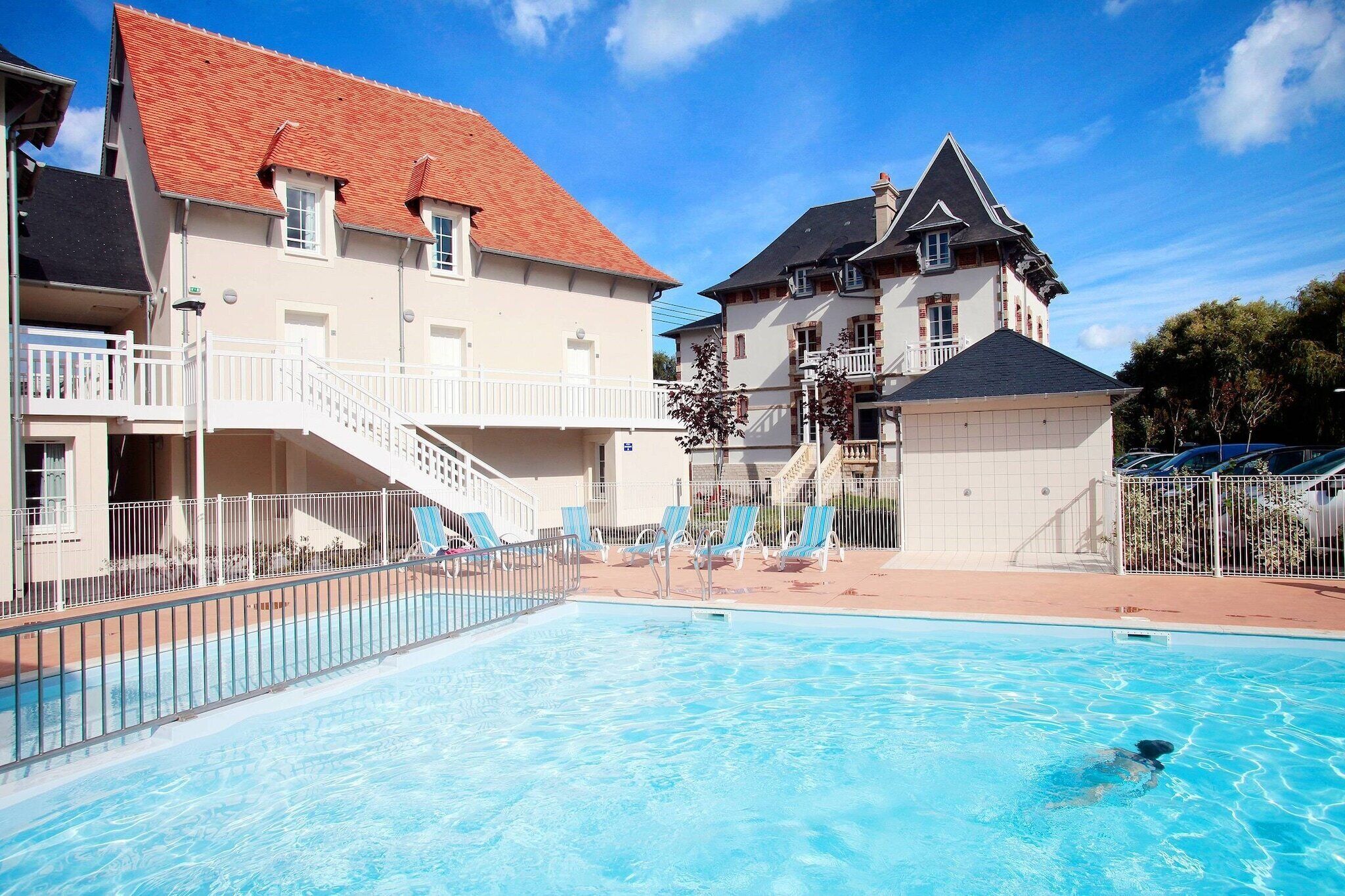 A swimming pool with lounge chairs, a building with a red roof, and a staircase leading to a balcony.