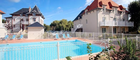 A swimming pool with lounge chairs, surrounded by a white fence, and adjacent to a building with a red roof.
