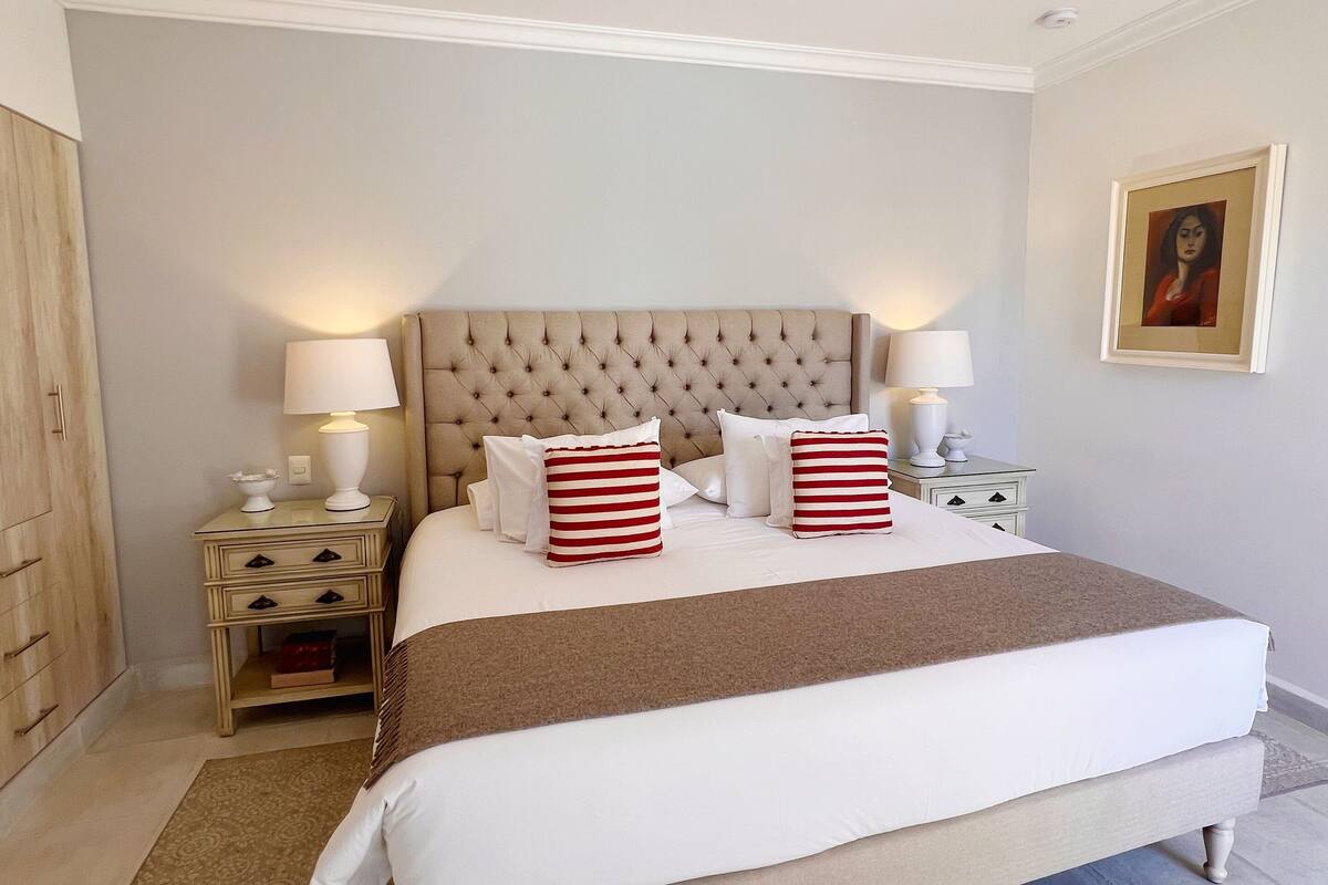 A neatly arranged bedroom with a large bed, two bedside tables with lamps, a headboard, and a framed picture on the wall.