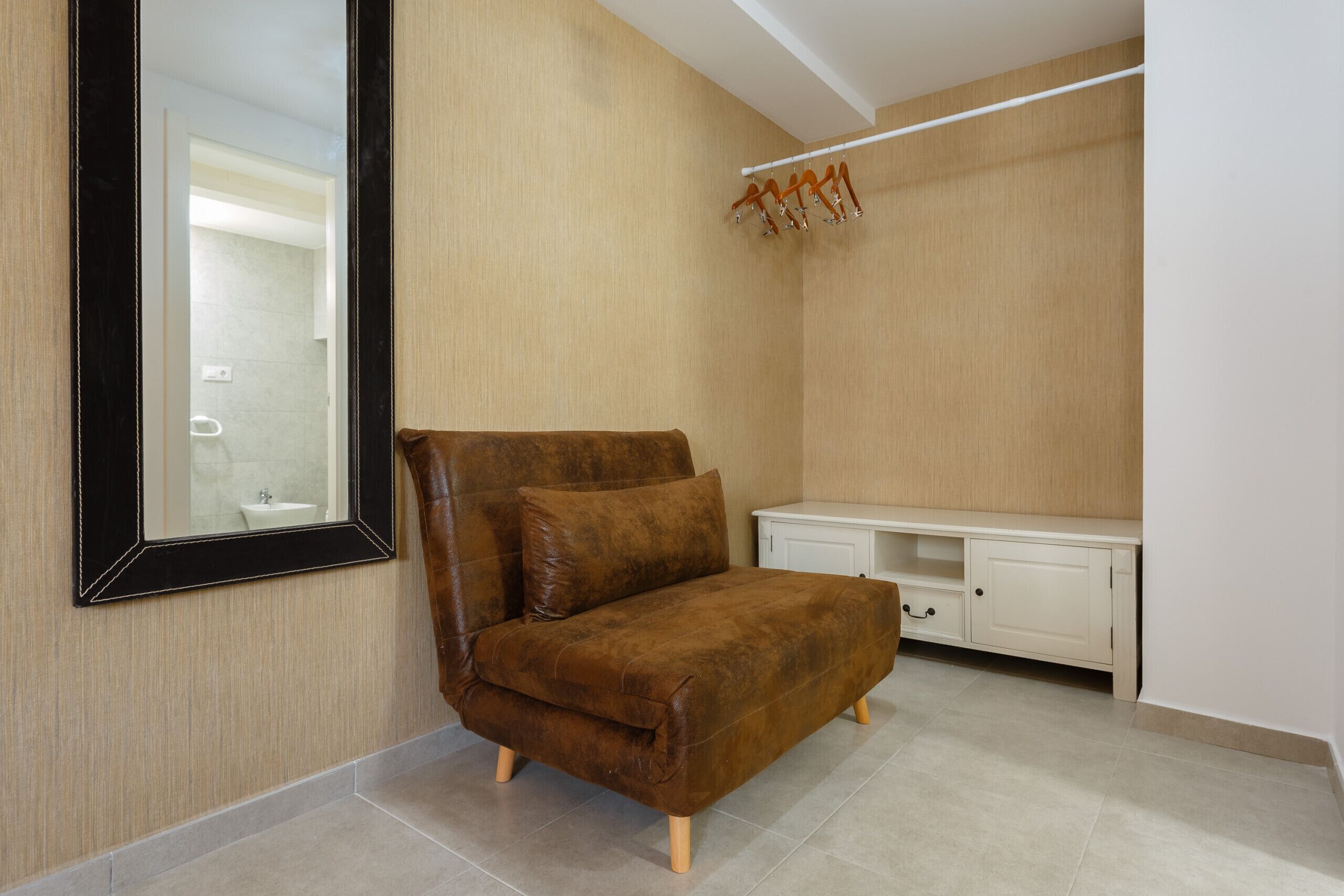 Un canapé en cuir marron dans une pièce où se trouvent un grand miroir, une armoire blanche et un banc en bois.