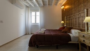 A bedroom with a bed, bedside lamps, a wooden headboard, a window with curtains, and an air conditioning unit. - Atico del ROMANTICO Gaditano by Cadiz4Rentals (Cádiz)