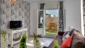 A living room with a flat-screen TV on a white console, a brown sofa with colorful cushions, and a view of an outdoor patio with a table and chairs. - Atico del ROMANTICO Gaditano by Cadiz4Rentals (Cádiz)