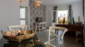 A dining area with a glass table, two white chairs, a woven basket with fruits, a small white cabinet with a television, and a view of a courtyard with plants and a wooden bench. - Atico del ROMANTICO Gaditano by Cadiz4Rentals (Cádiz)