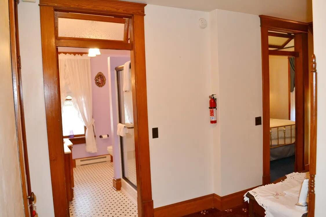 A hotel room with a wooden door, a white wall, a bed with a white bedspread, and a small table with a white tablecloth.