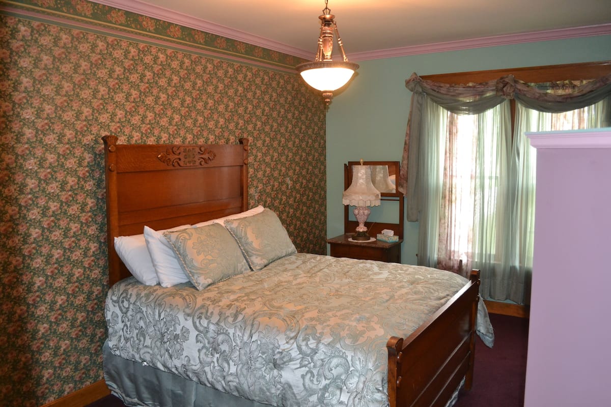 A bedroom with floral wallpaper, a wooden bed, a nightstand with a lamp, and a window with curtains.
