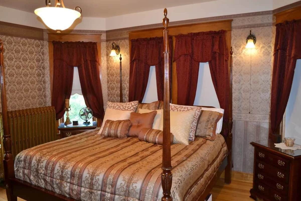 A four-poster bed with a wooden frame and a striped bedspread, flanked by two nightstands with lamps, in a room with patterned wallpaper and red curtains.