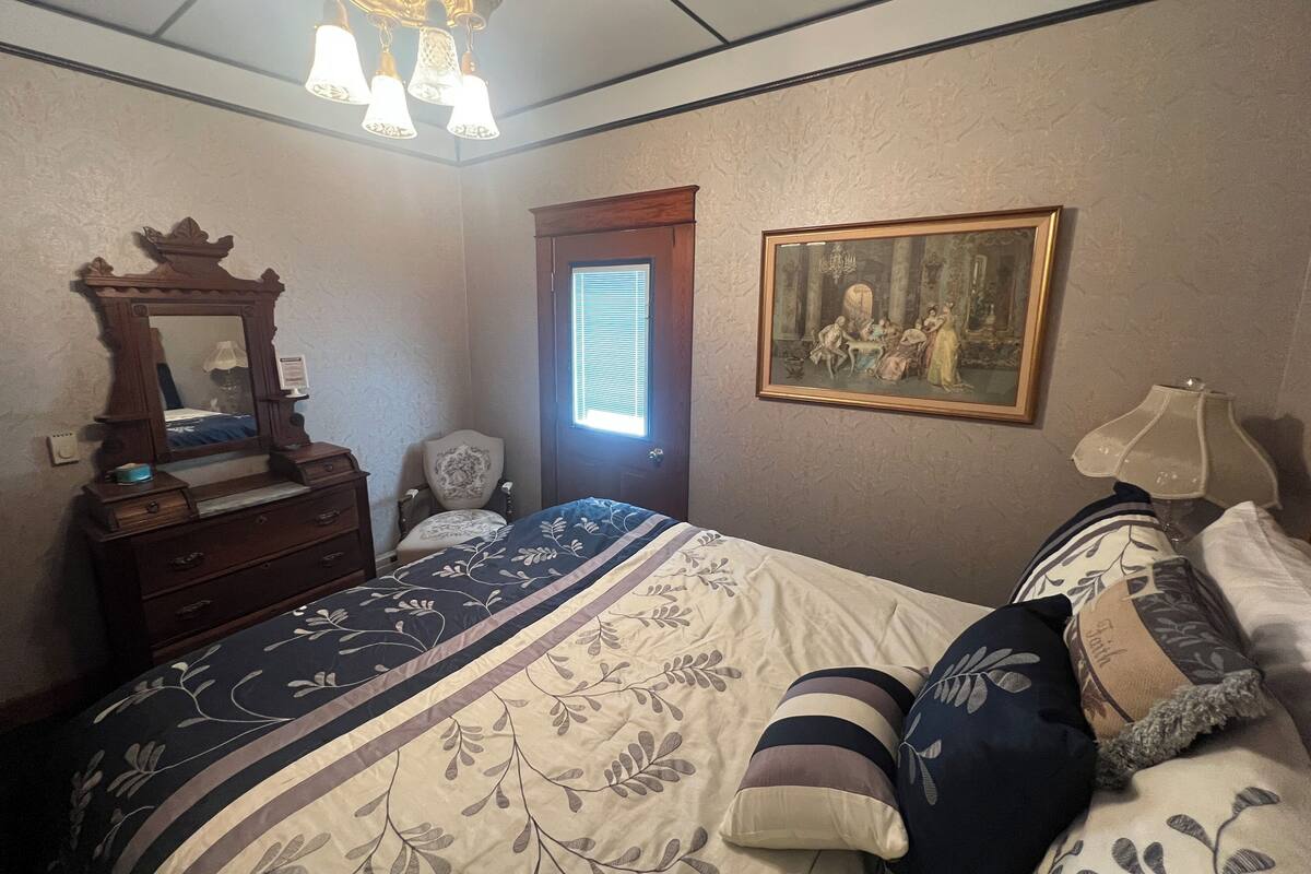 A bedroom with a bed, a dresser, a mirror, a chair, a painting, and a window.