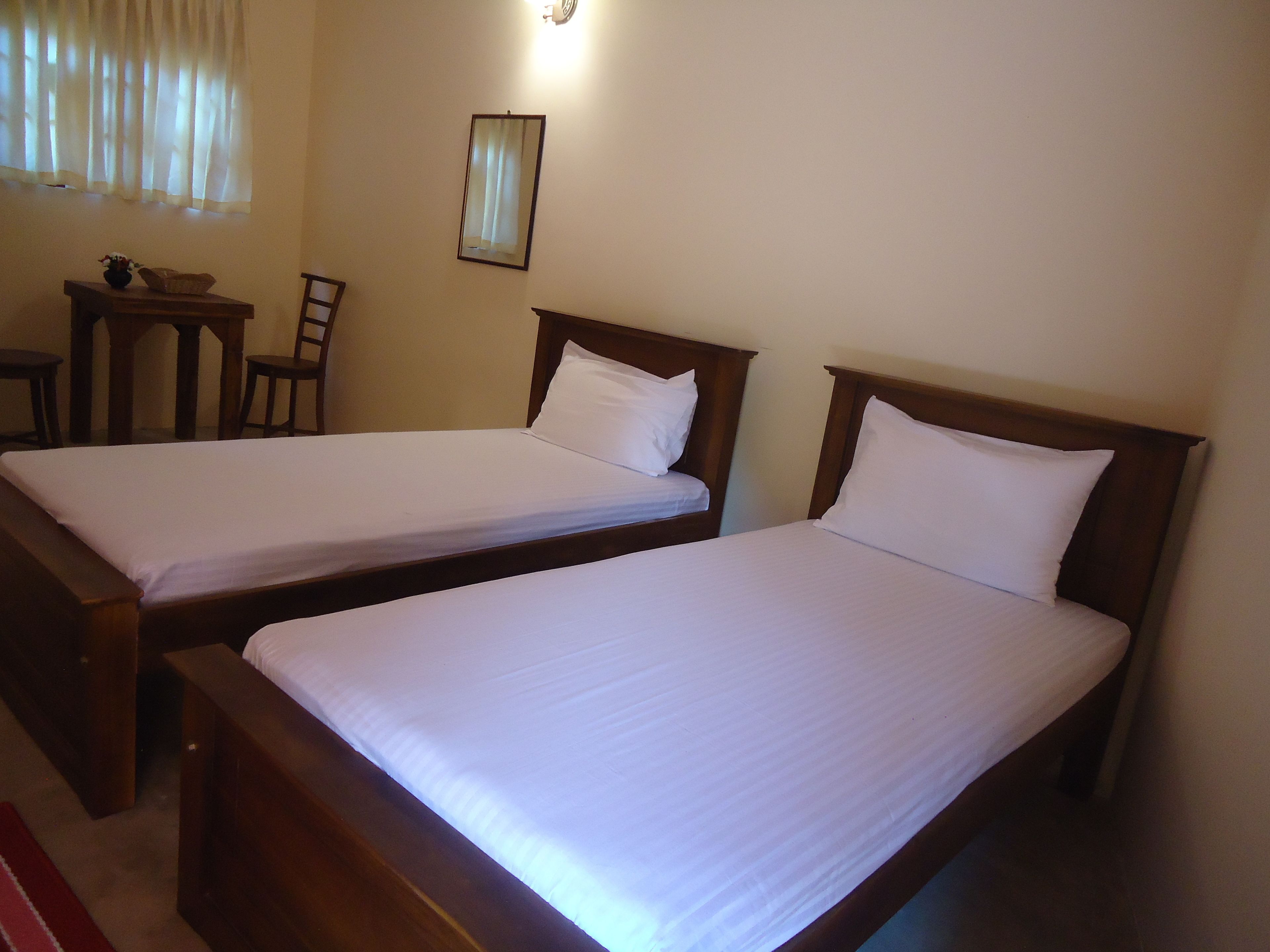Two single beds with wooden frames and white bedding in a room with a small table and chair, a mirror on the wall, and a window with sheer curtains.