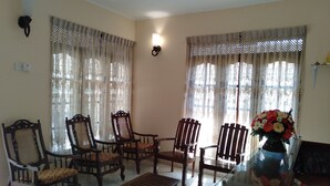 Una habitación con cuatro sillas de madera con cojines estampados, una mesa de centro de vidrio y dos ventanas con cortinas decoradas.
