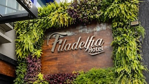 En hotellentrén med en skylt som visar texten ”Filadelfia Hotel”, omgiven av frodig grönska och en entré med svarta, stängda luckor.