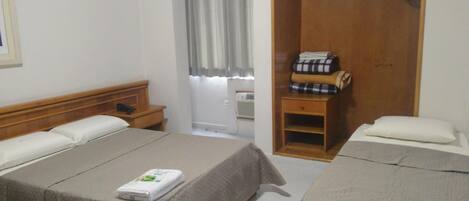 A hotel room with two beds, a wooden wardrobe, a small table with folded clothes, and a window with curtains.