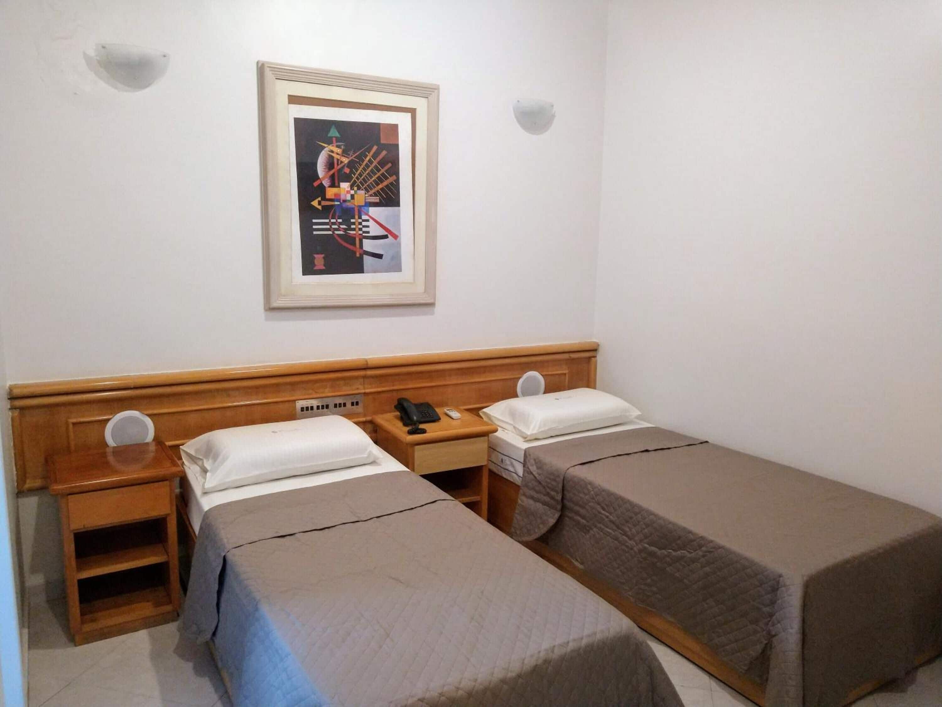 A hotel room with two single beds, a wooden headboard, a bedside table with a phone and a remote control, and a framed picture on the wall.