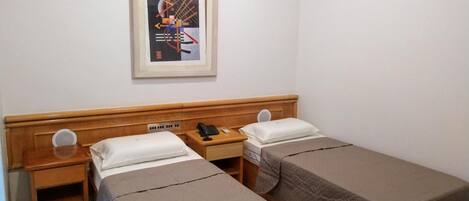 A hotel room with two single beds, a wooden headboard, a bedside table with a phone and a remote control, and a framed picture on the wall.
