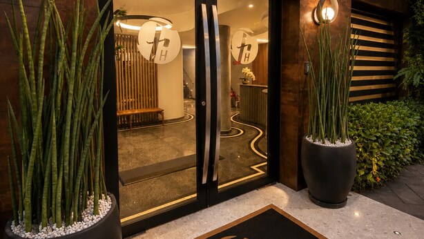 En hotellentrén med modern design, med en glasdörr, en dörrmatta med hotellets namn och två stora krukväxter.