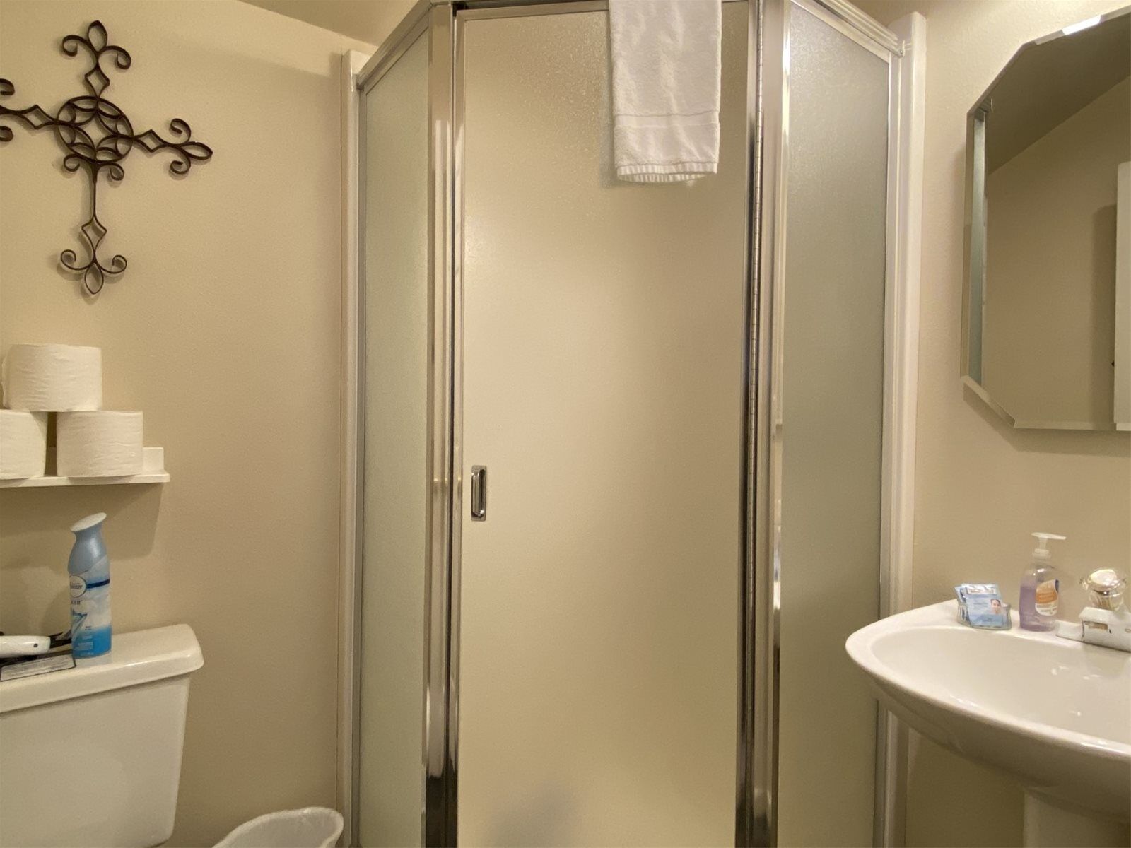 Um banheiro com box de vidro, vaso sanitário com papel higiênico e uma garrafa azul sobre a caixa, pia com dosadores de sabonete e uma toalha pendurada na porta do chuveiro.