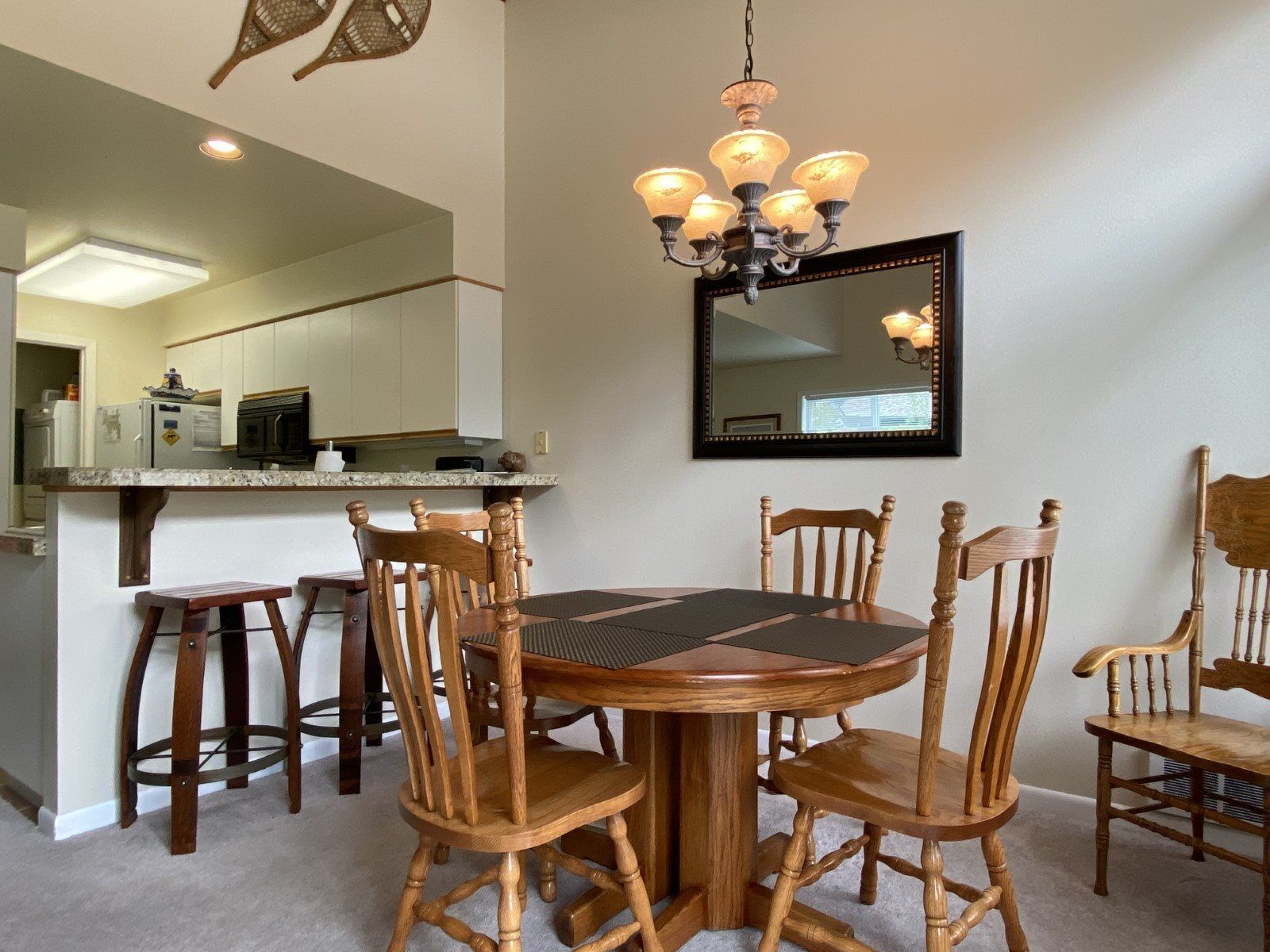 A dining area with a round wooden table surrounded by six wooden chairs. A chandelier hangs above the table, and a large mirror is mounted on the wall.