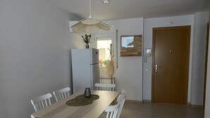 Un coin repas avec une table en bois, des chaises blanches, un luminaire suspendu en fibres tressées, un réfrigérateur et une porte en bois.