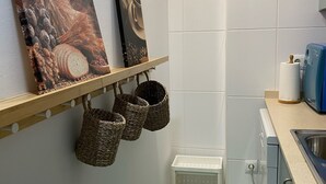 Une petite cuisine avec des carreaux blancs, un évier, un lave-vaisselle et un plan de travail sur lequel se trouve un appareil électroménager bleu. On y trouve également deux paniers fixés au mur et un petit tabouret blanc.