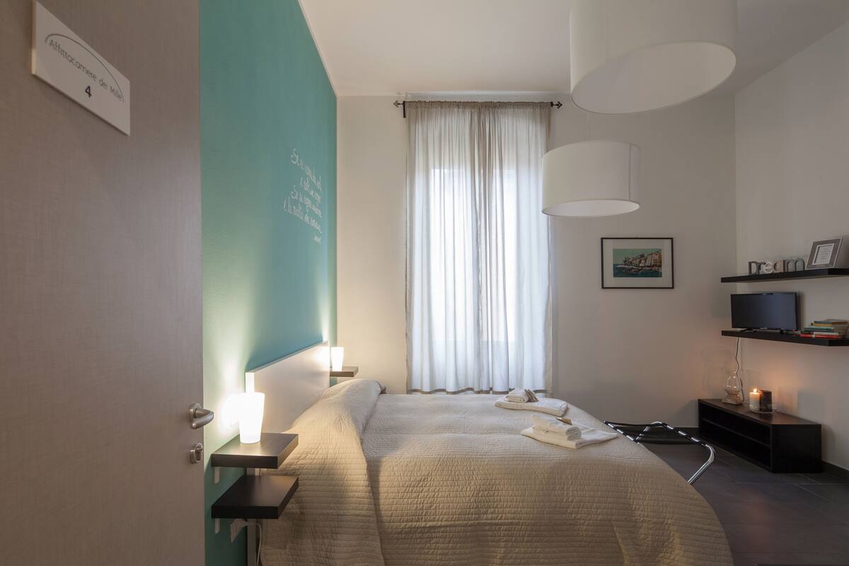 Habitació d'hotel amb llit, taulells laterals, televisió a una estant i finestra amb cortines primes.