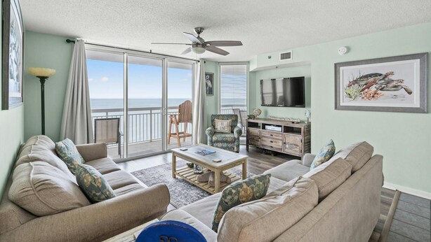 A living room with a sofa, coffee table, and a TV. There's a balcony with a view of the ocean. - Luxury 15th Floor Corner Unit, Jacuzzi Tub, Lazy River | Crescent Shores - S 1506 (North Myrtle Beach)