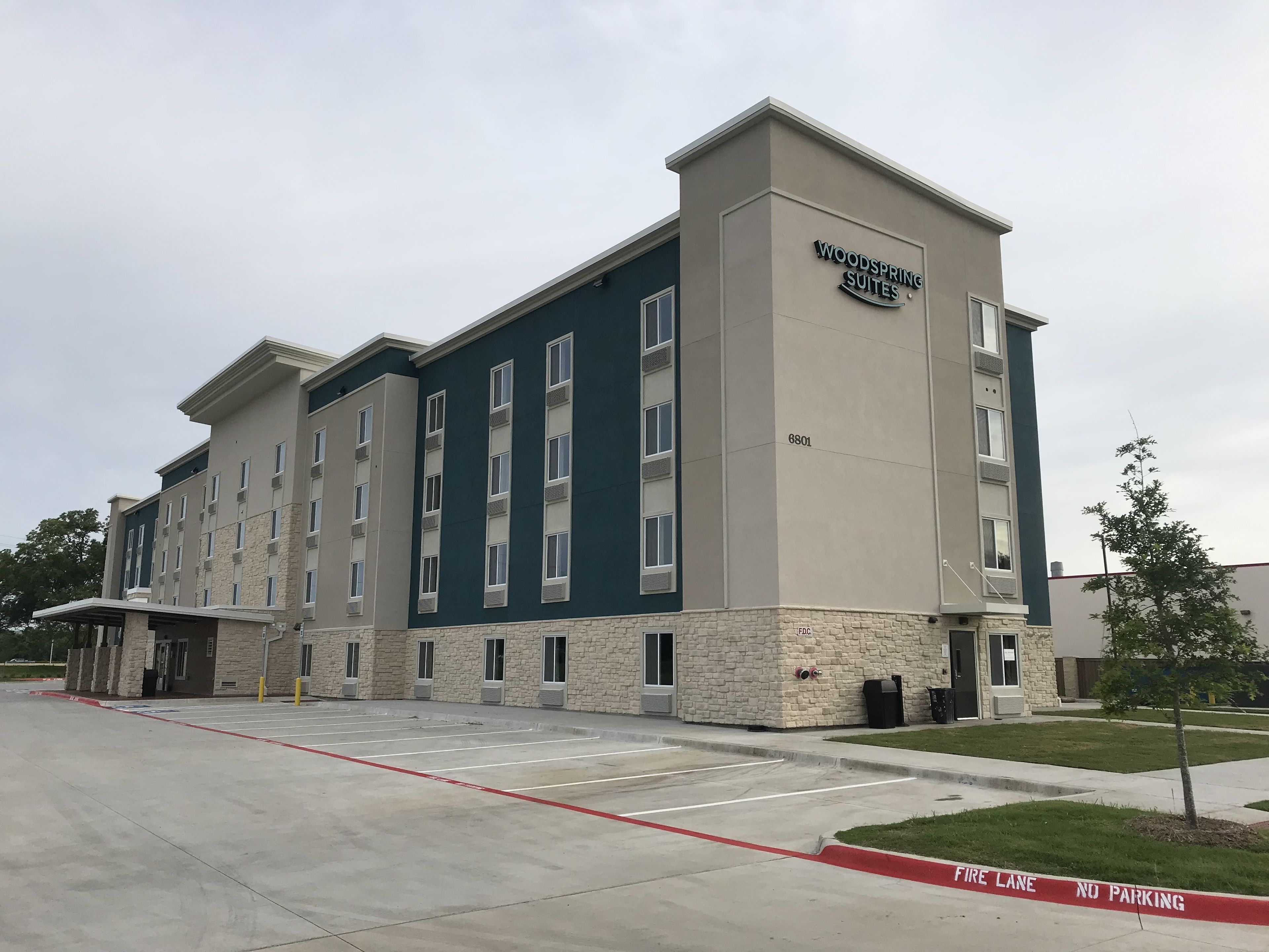 A multi-story hotel with a sign reading "Woodspring Suites." It features a modern architectural design with a combination of stone and painted exterior walls.