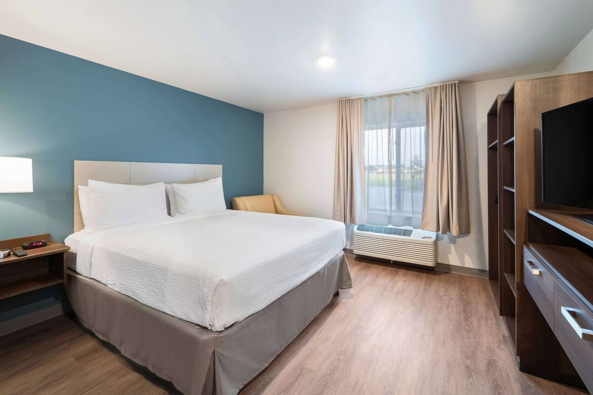 A hotel room with a bed, a nightstand, a TV, a window with curtains, and a built-in air conditioning unit.