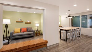 A modern living room with a sofa, a wooden bench, a dining table with chairs, and a small side table with a lamp. - WoodSpring Suites Dallas Plano Central Legacy Drive (Plano)