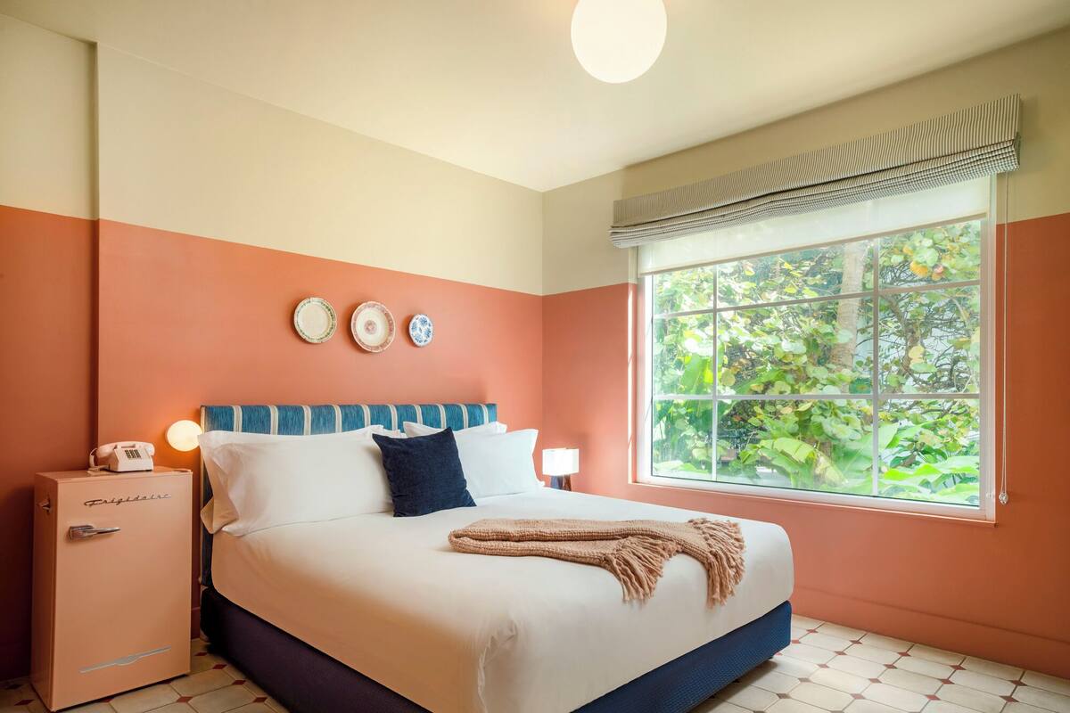 A bedroom with a bed, bedside table, lamp, and a window with a view of greenery.