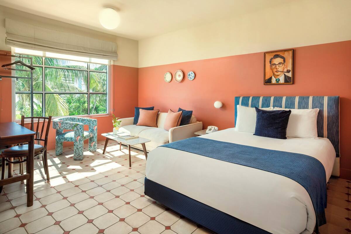 A hotel room with a bed, a desk, a chair, a window with blinds, a painting on the wall, and a tiled floor.