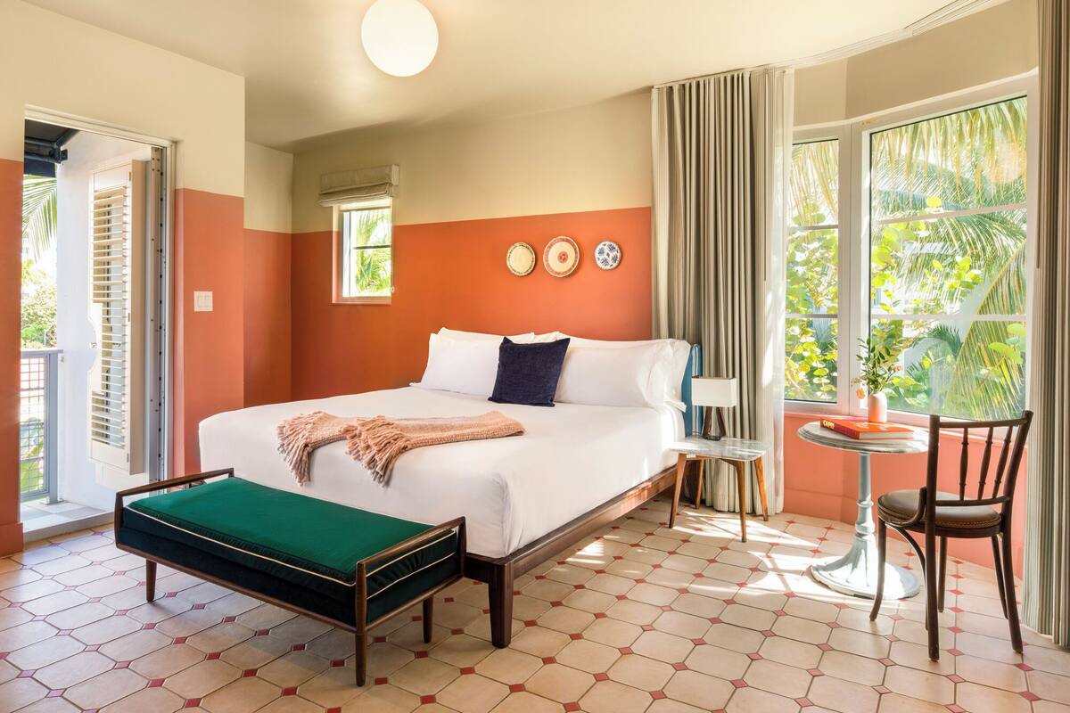 A hotel room with a bed, a small table, a chair, a window with curtains, and a view of greenery outside.
