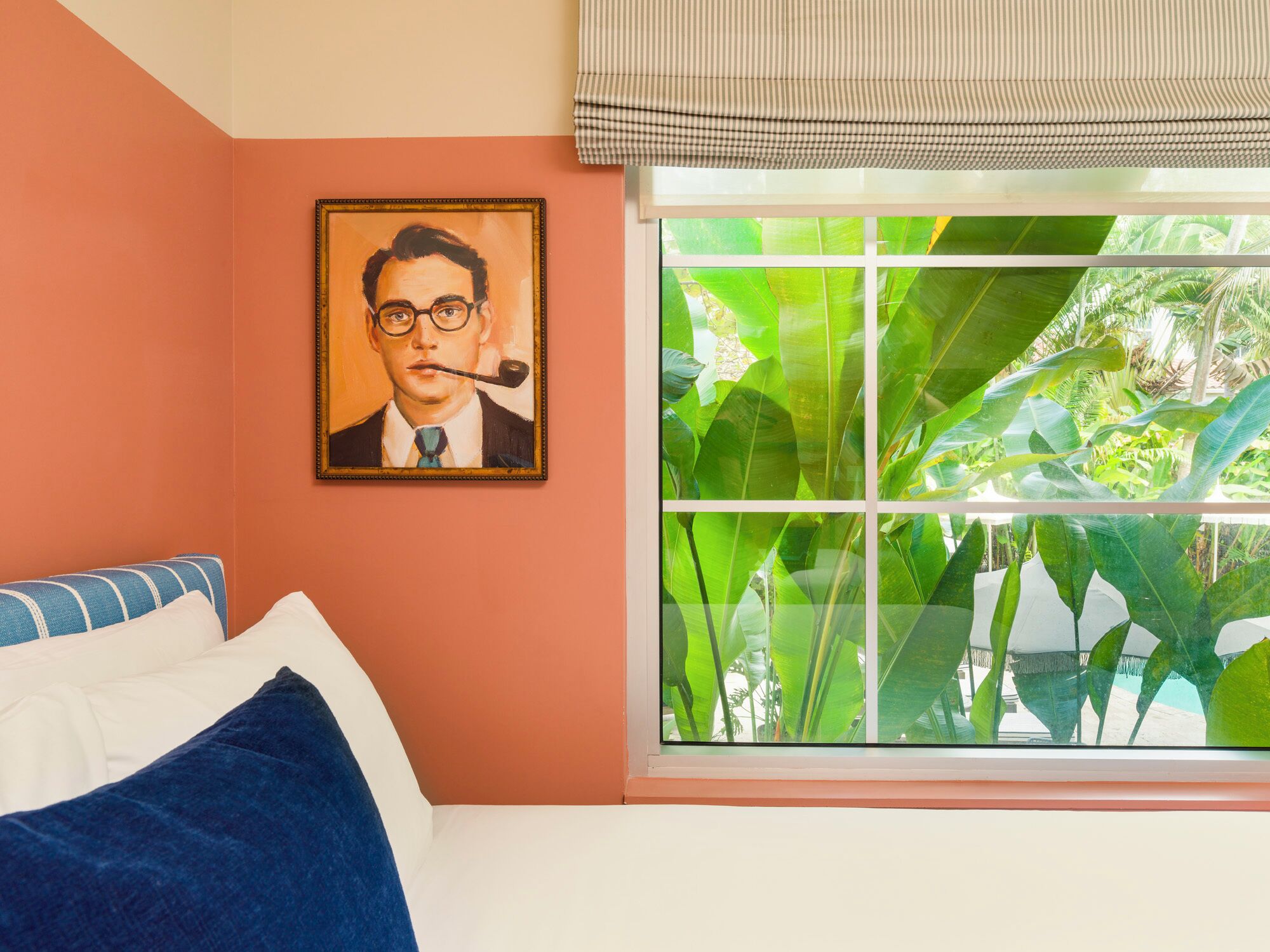 A bedroom with a bed, pillows, a painting, a window with a view of greenery, and a striped curtain.