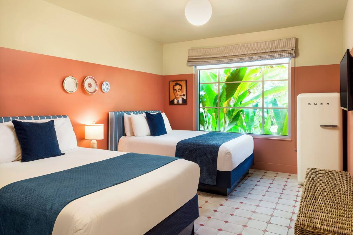 A hotel room with two beds, a window with a view of greenery, a wall-mounted plate, a framed picture, and a white refrigerator.