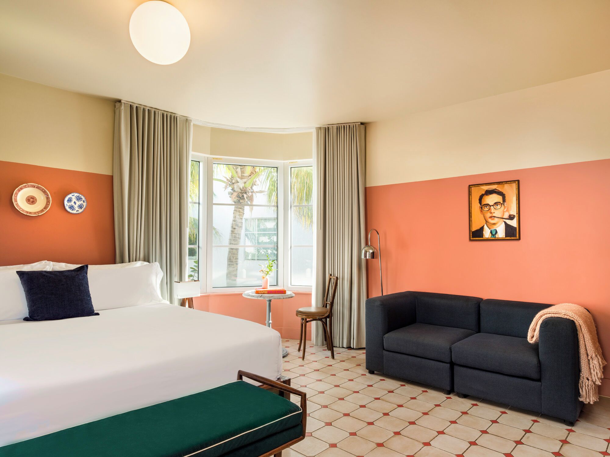 A hotel room with a bed, a sofa, a small table, a chair, a painting, and a window with curtains.
