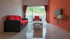 A room with a sofa, a chair, a small table, a TV on a shelf, and a view of greenery outside. - Cameron Highlands (Cameron Jaya) (Cameron Highlands)
