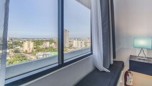 A hotel room with a large window offering a city view, a bedside table with a lamp, and a fire extinguisher on the wall. - Joseph's Place @ Avenir Condo w/ NETFLIX (Cebu City)