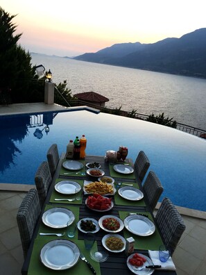 A dining table set for a meal by a pool with a view of the sea and mountains. - Villa Mavi Manzara / Luxury, secluded private villa (Kas)