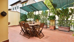 Een terras met een houten tafel en stoelen, omringd door plantenpotten en een groen bladerdak.